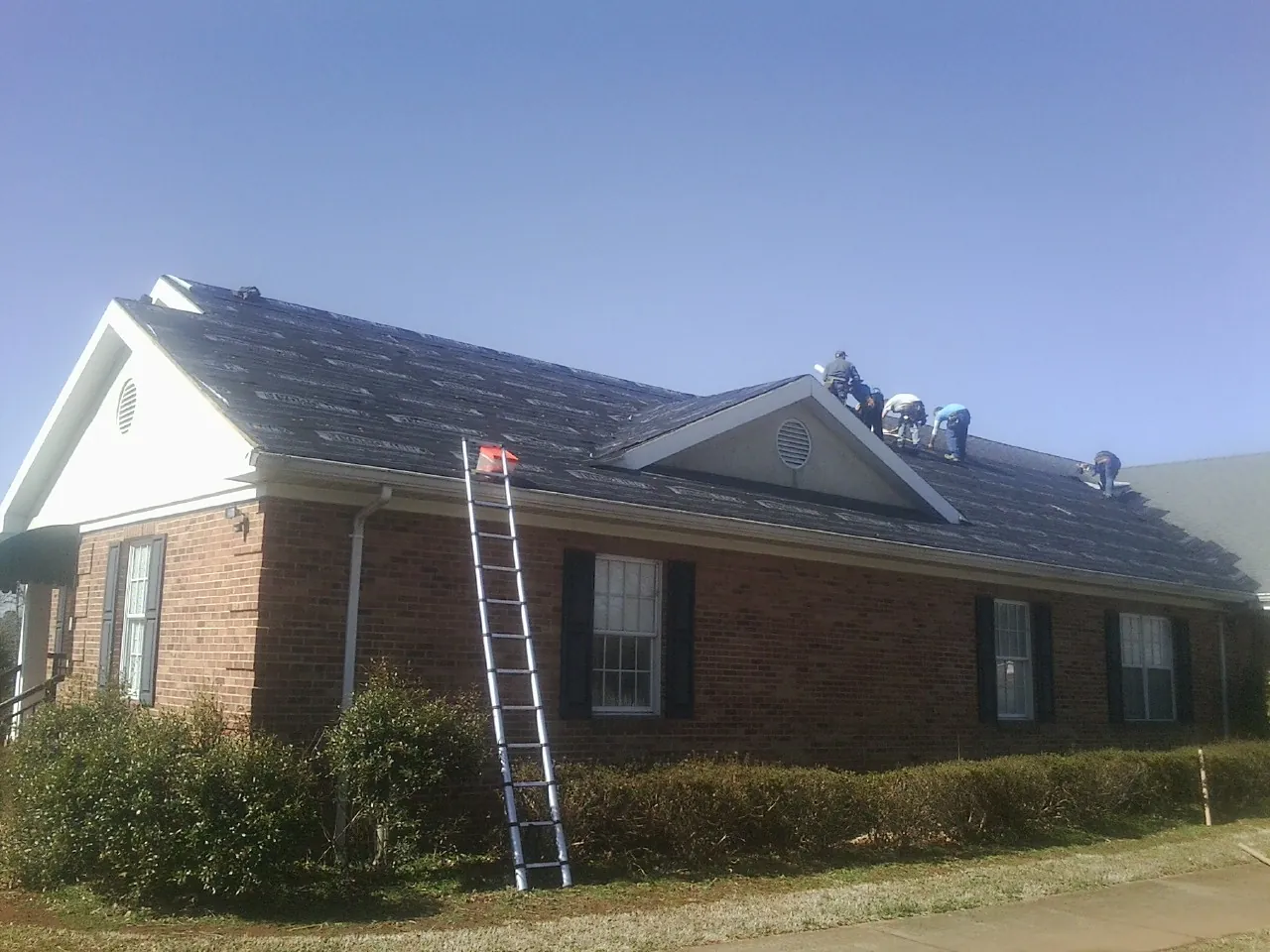 Crew tearing shingles off a brick ranch roof with a ladder set — Metal Roof Repair work by our crew in Casco