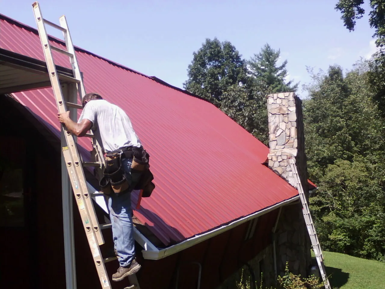 Roofer on a ladder beside a red metal roof and stone chimney — Soffit & Fascia Repair by Roofing Brothers in Casco